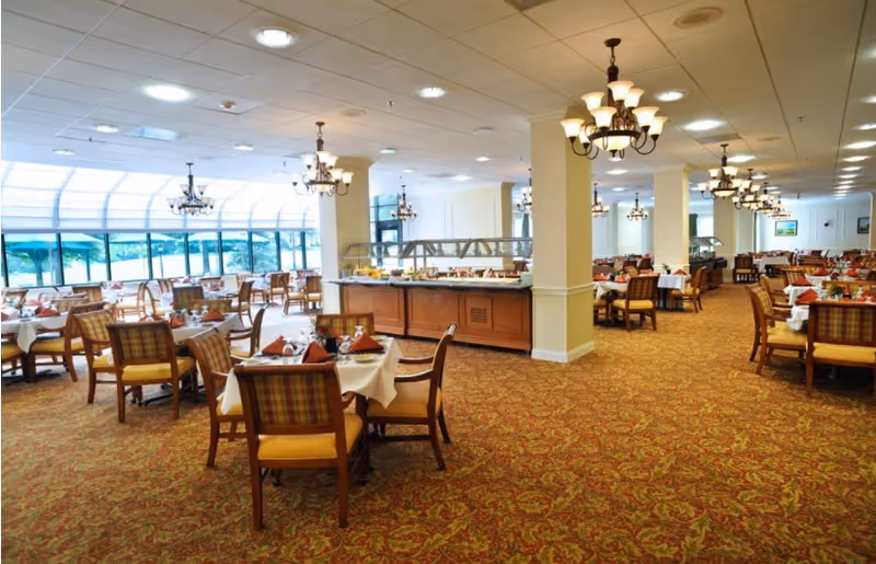 Spacious dining room with set tables, buffet stations, chandeliers, and large windows overlooking greenery.