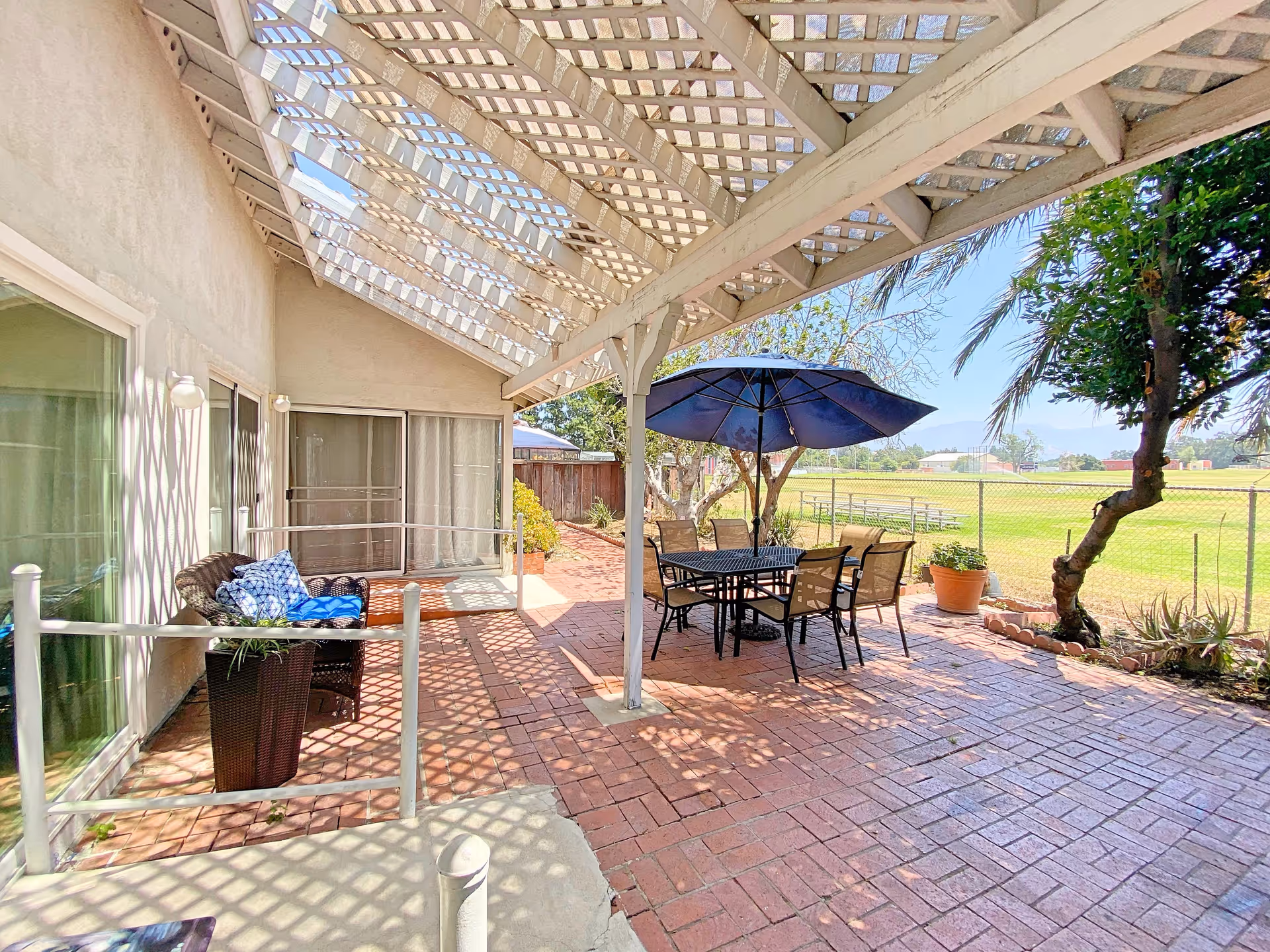 Outdoor patio area with a lattice roof casting shadows on the brick floor. There is a black metal table with six chairs and a large blue umbrella. To the left, there is a wicker loveseat with blue cushions and a potted plant. The patio overlooks a fenced grassy field with trees and mountains in the background.
