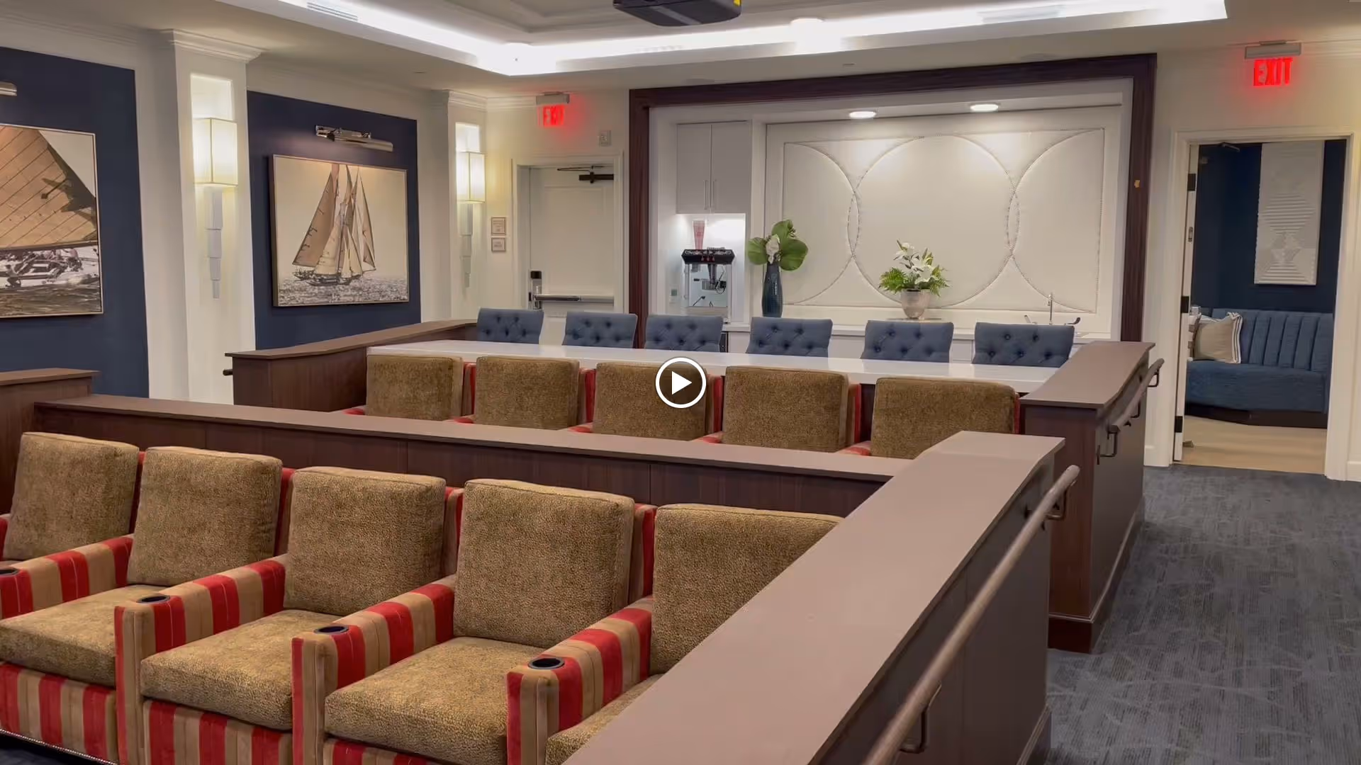 A well-lit interior room with multiple rows of comfortable chairs arranged in a tiered seating layout, featuring brown and red striped upholstery in the front row and blue tufted chairs in the back row. The walls are decorated with framed pictures of sailboats and elegant wall sconces. There is a water dispenser and floral arrangements on a counter at the back of the room, with doorways on either side leading to other rooms.