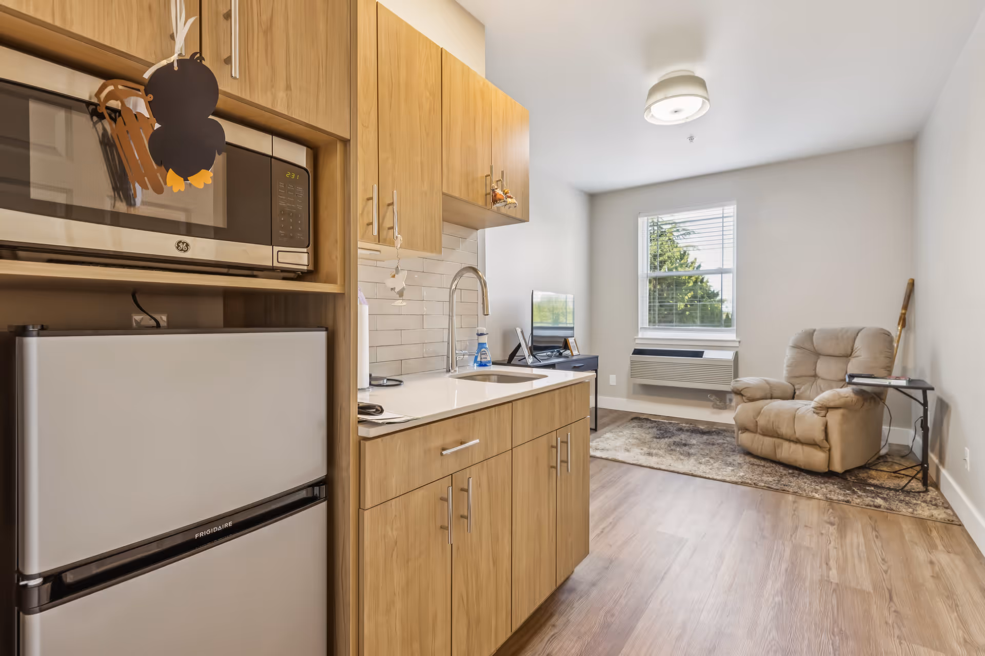 A small living space featuring a kitchenette with wooden cabinets, a microwave, and a mini refrigerator on the left. The kitchenette has a white countertop with a sink and a tiled backsplash. To the right, there is a cozy beige recliner chair on a patterned rug near a window with white blinds. A small black side table is next to the chair, and a flat-screen TV is placed on a black stand against the far wall. The room has light wood flooring and white walls with a ceiling light fixture.