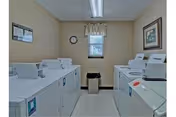 Small laundry room with multiple washers and dryers, a window, wall clock, and framed artwork.