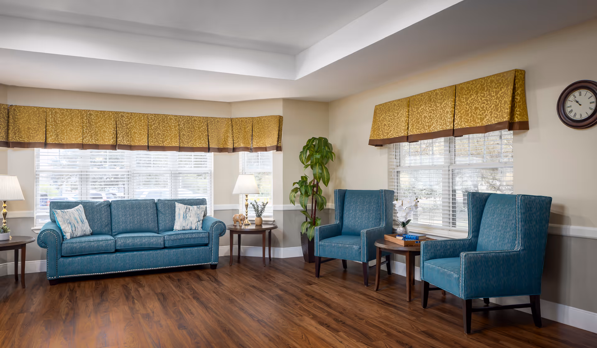 A bright and spacious living room area with wooden flooring, featuring a teal upholstered sofa with two patterned pillows, two matching teal armchairs, two round wooden side tables with lamps and decorative items, large windows with yellow and brown valances, a tall green plant in the corner, and a wall clock.