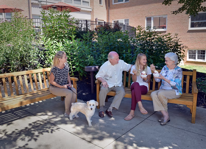 Four people sitting on wooden benches outside near a garden area. Two women and one man are seated on one bench, with the man in the middle. Another woman is seated on a separate bench holding a small dog on a leash. Two of the women are eating from bowls, possibly ice cream or dessert. The setting is sunny with shadows from nearby trees and a brick building in the background.