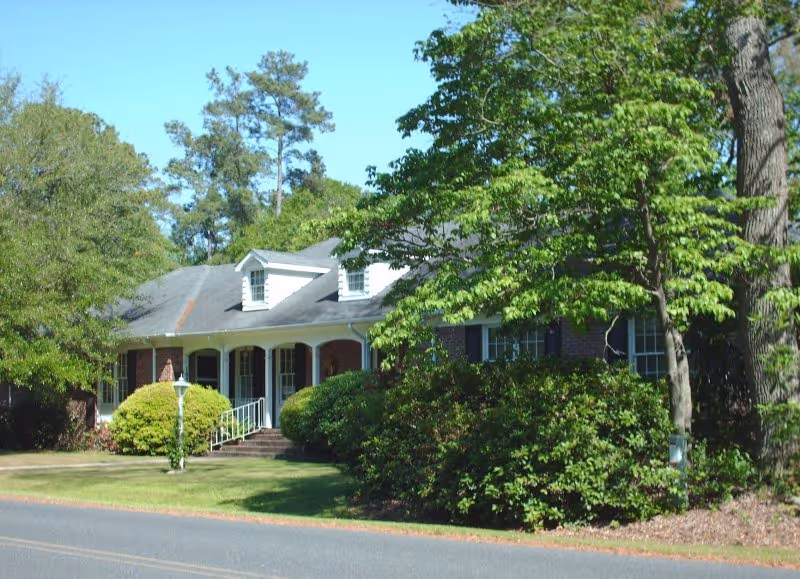 Exterior view of a single-story brick building with a gray shingled roof, white columns, and a small porch. The building is surrounded by green trees and bushes under a clear blue sky.