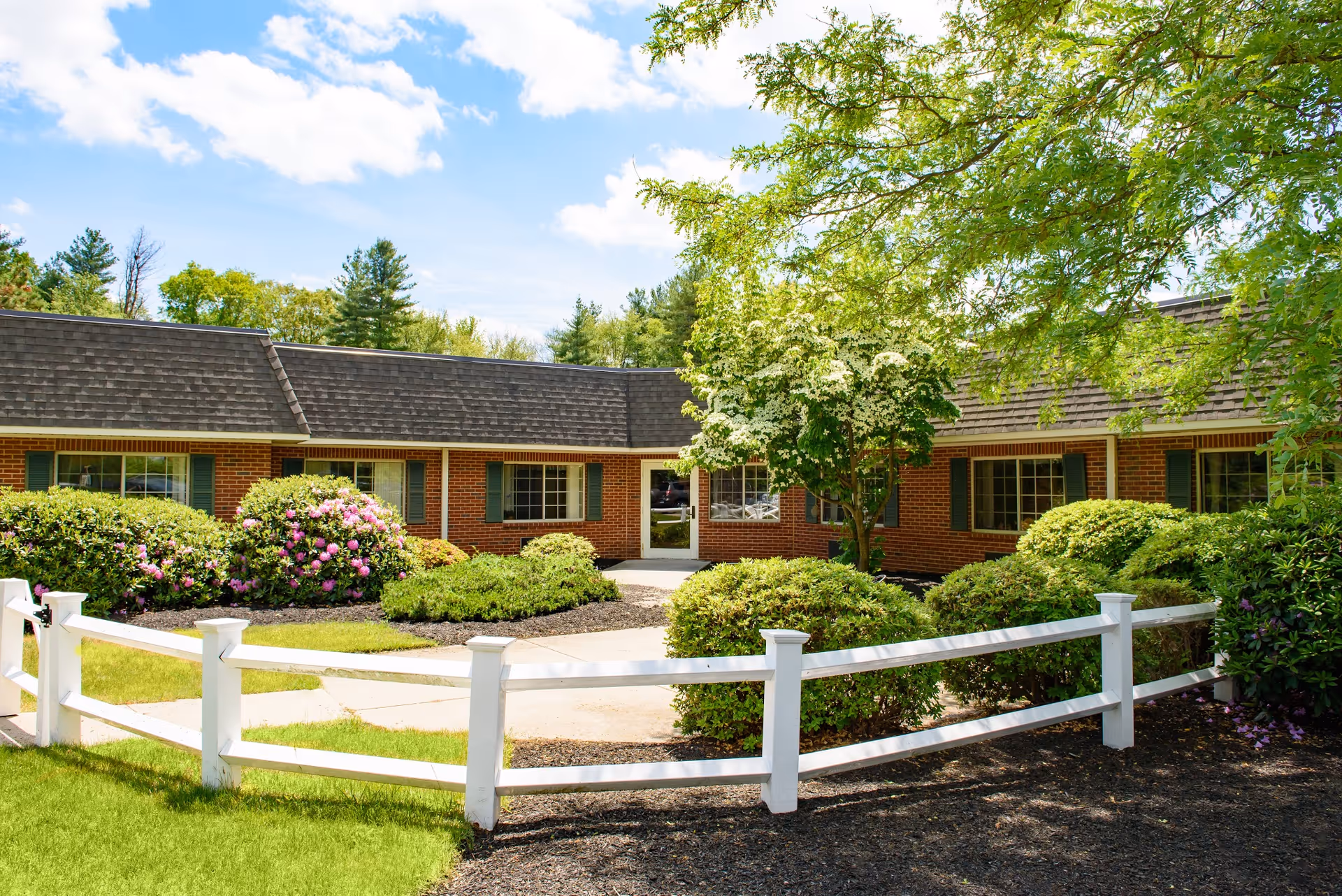 Front entrance of a single-story brick senior living building with a white fence, landscaped shrubs and trees under a blue sky.