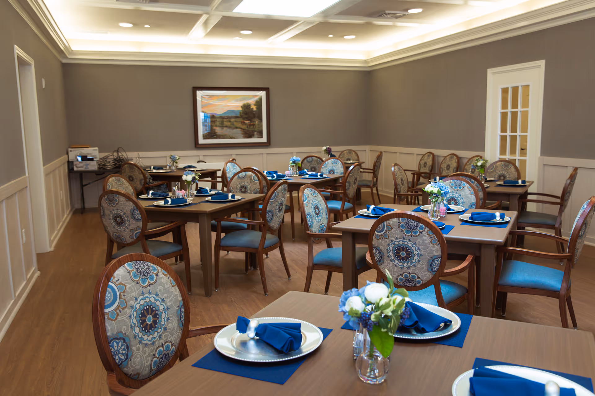 A dining room with multiple wooden tables and chairs featuring blue and patterned upholstery. Each table is set with plates, blue napkins, and small floral centerpieces. The room has wooden flooring, gray walls with white wainscoting, recessed ceiling lights, and a framed landscape painting on the wall.