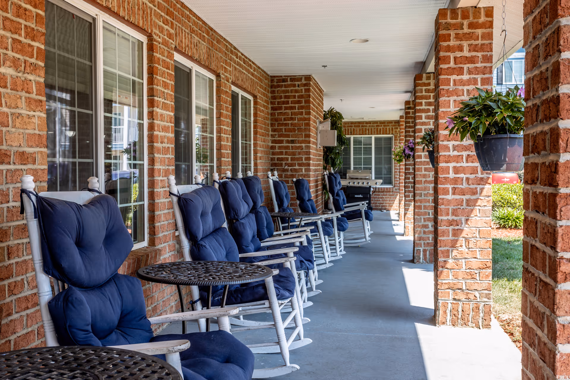 A covered outdoor patio area with a row of white rocking chairs with blue cushions lined up against a brick wall with windows. There are small round metal tables in front of the chairs and hanging flower pots attached to the brick columns. A grill is visible in the background near the end of the patio.