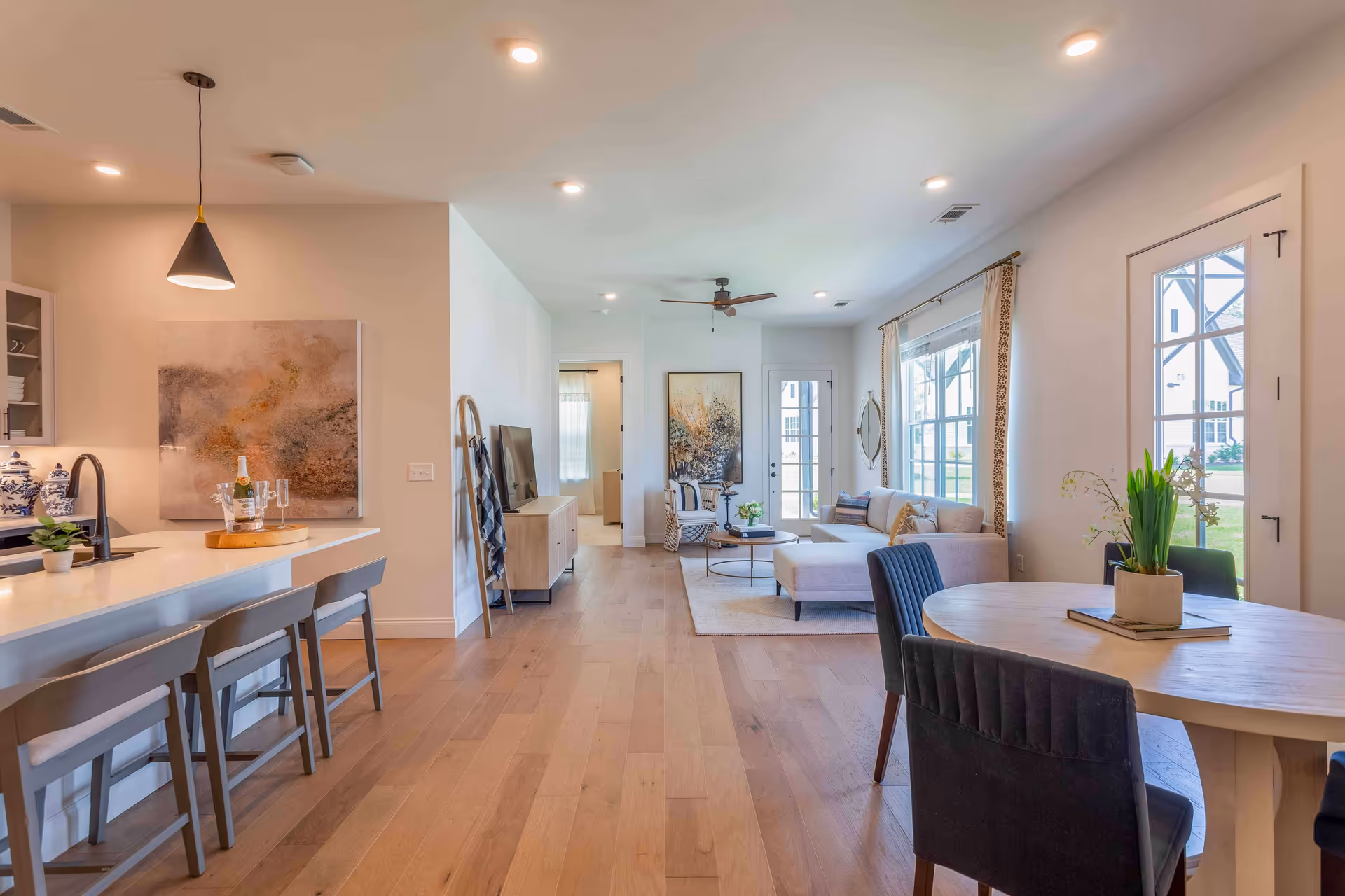 Bright and modern open-concept living space with light wood flooring, a kitchen island with three gray bar stools, a round dining table with four dark upholstered chairs, and a cozy living room area featuring a light gray sectional sofa, a round coffee table, and a ceiling fan. Large windows and glass doors allow natural light to fill the room, and decorative artwork and plants add warmth to the space.