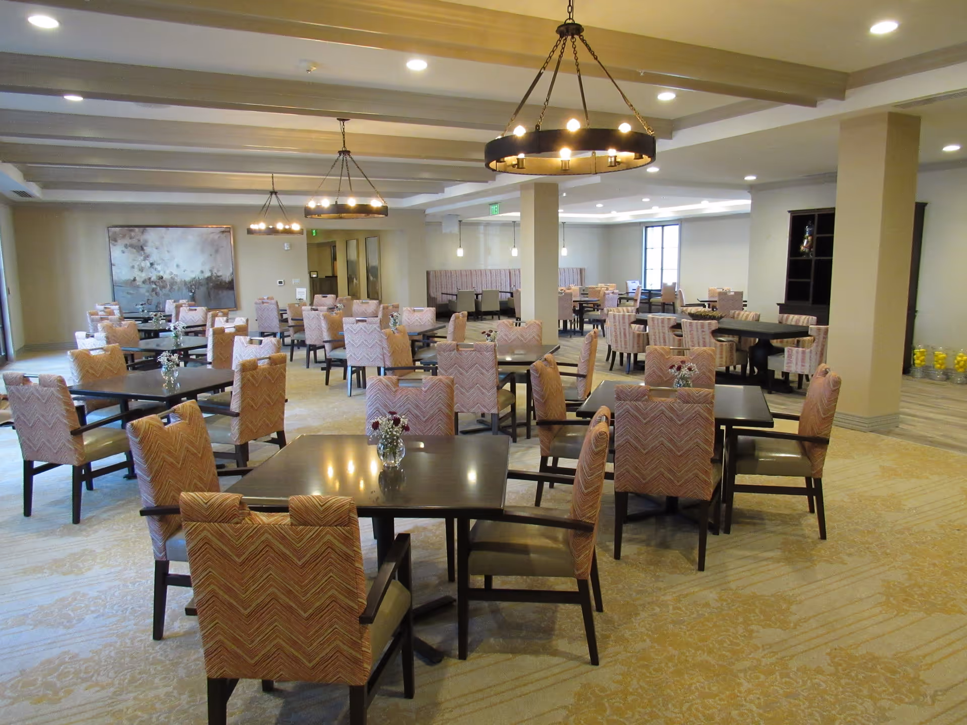 A spacious dining room with multiple square tables and cushioned chairs arranged neatly. The room features beige walls, patterned carpet, modern chandeliers hanging from the ceiling, and a large abstract painting on one wall. There are small flower arrangements on each table, and natural light enters through windows in the background.