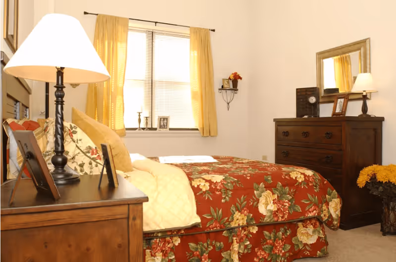 A cozy bedroom with a bed covered in a floral red, yellow, and green bedspread. There is a wooden nightstand with a lamp and picture frames on the left side of the bed. A window with yellow curtains is behind the bed. On the right side of the room, there is a wooden dresser with a mirror, a lamp, and decorative items. A small shelf with a flower vase is mounted on the wall above the dresser.