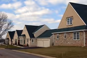 Exterior view of cottage-style senior living buildings with garages, pitched roofs, and a grassy front lawn under a partly cloudy sky.