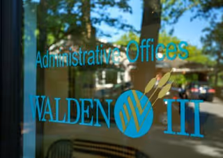 Glass door with blue text that reads 'Administrative Offices WALDEN III' with a reflection of trees, cars, and a building in the background.