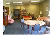 A common area in a senior living facility featuring a round table with four chairs in the foreground, a pink armchair, a bookshelf, a television, a pool table, a sofa, and a floor lamp. The room has carpeted flooring and neutral-colored walls with framed artwork.