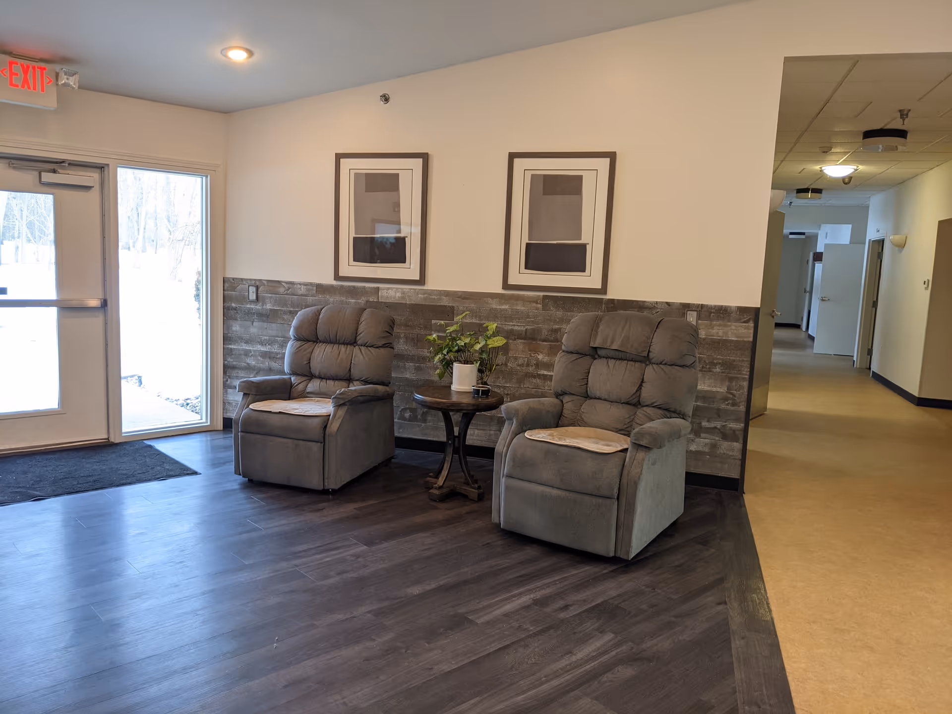 A seating area inside a facility with two gray recliner chairs separated by a small round wooden table holding a potted plant. The wall behind the chairs has a wooden panel halfway up and two framed abstract artworks. To the left is a glass door with an exit sign above it, and to the right is a hallway with several open doors.