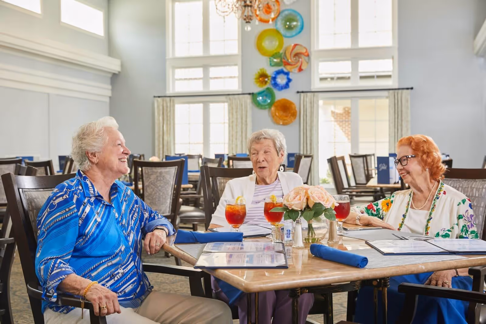 Three elderly women sitting around a dining table in a bright room with large windows and colorful wall decorations, enjoying drinks and conversation.