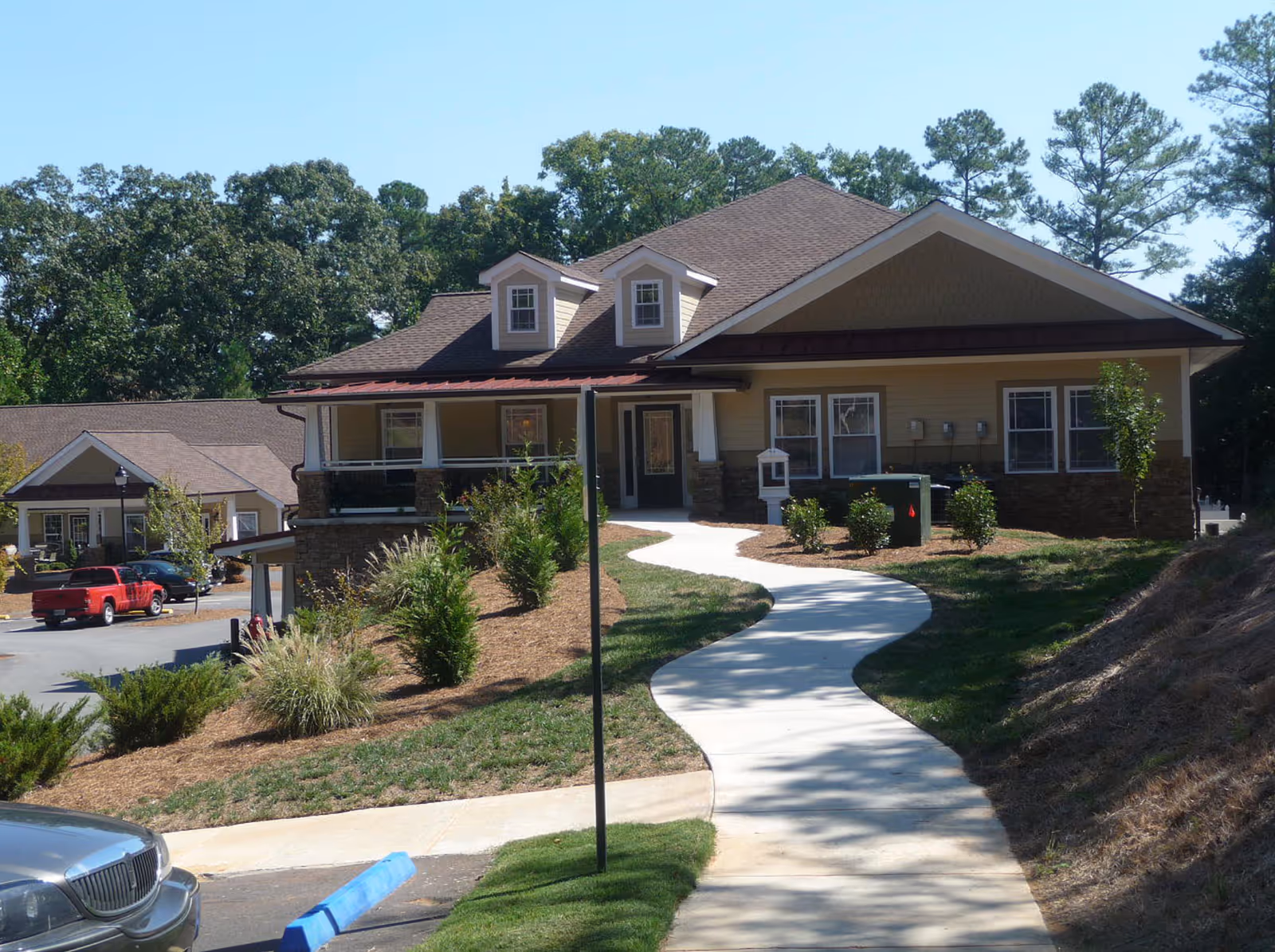 A single-story building with a brown roof and beige siding, surrounded by landscaped bushes and trees. A curved concrete walkway leads up to the front entrance. There are other buildings and parked vehicles visible in the background under a clear blue sky.