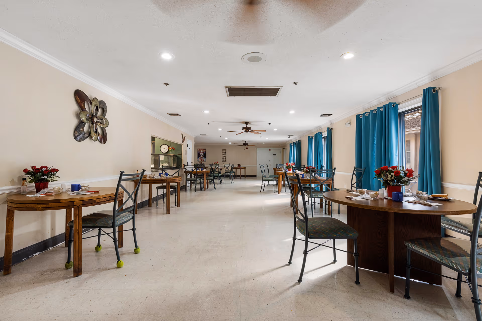 A spacious dining room with round and rectangular tables set for meals, metal chairs, blue curtains, and ceiling fans.