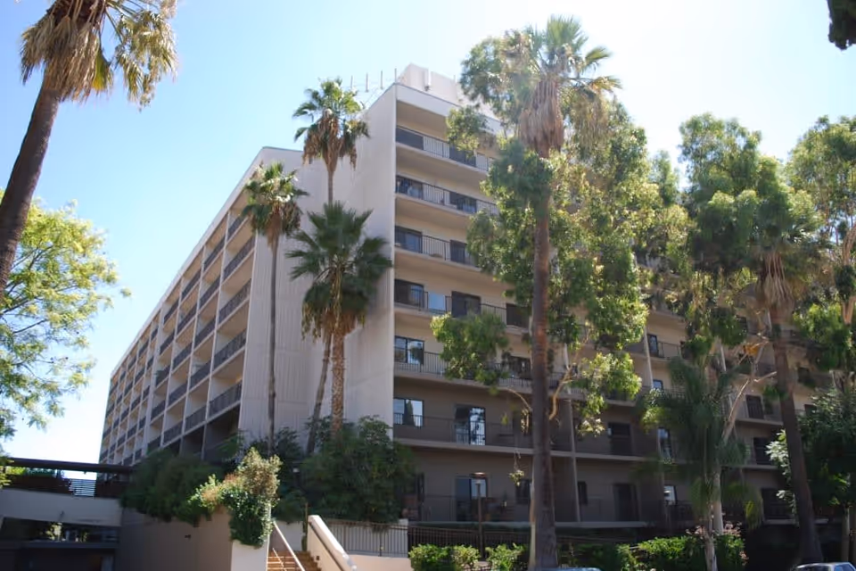 Exterior view of a multi-story senior living facility building surrounded by tall palm trees and other greenery under a clear blue sky.
