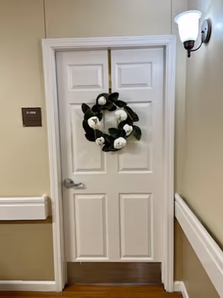 Closed white paneled door in a hallway adorned with a magnolia wreath and lit by a wall sconce.
