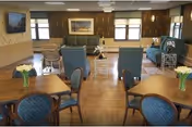 A spacious living room area in a senior living facility with wooden flooring, several tables with chairs, blue upholstered armchairs, and a green sofa. The room has large windows letting in natural light, a wall-mounted TV, and framed artwork on the walls. Small vases with white flowers are placed on the tables.