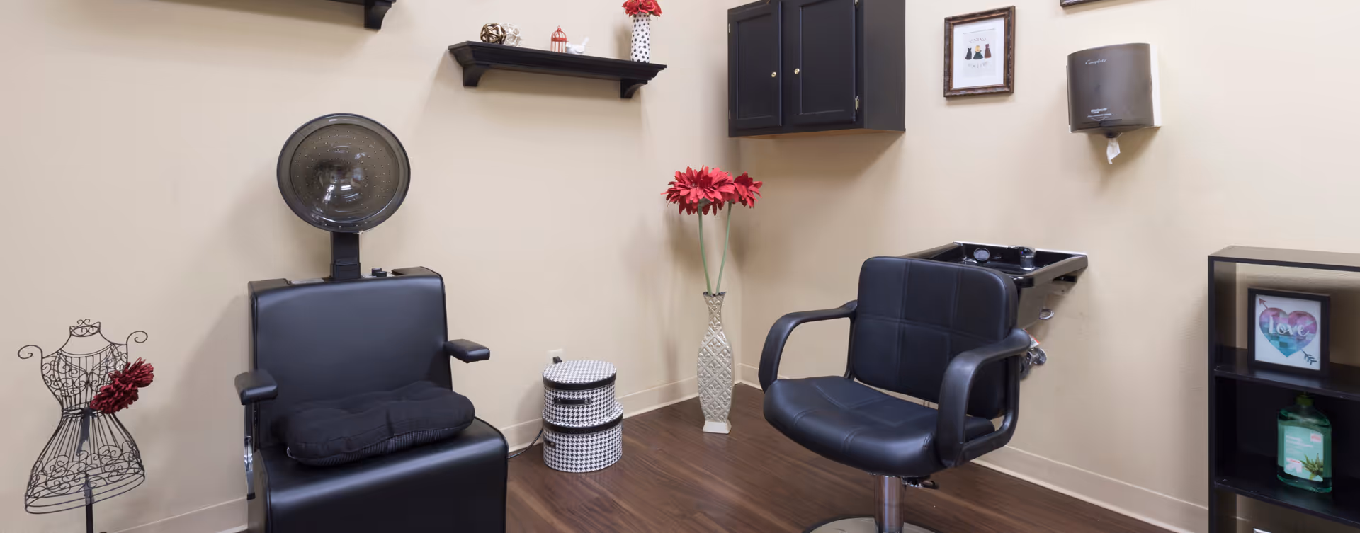 Small salon-style room with two black styling chairs, a hooded hair dryer, a wash sink, and decorative shelves and flowers.