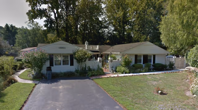 Single-story residential building with a light-colored exterior surrounded by greenery and trees. A paved driveway leads up to the entrance, which is framed by bushes and flowering plants. The building has a low-pitched roof and several windows with shutters.