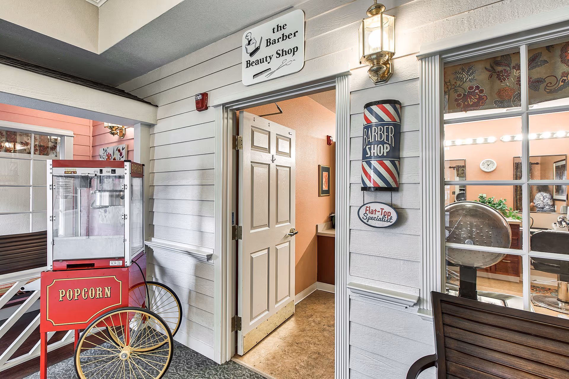 Entrance to a barber beauty shop inside a senior living facility, with a vintage-style popcorn machine on wheels nearby. The door to the barber shop is open, showing part of the interior with a sink and framed artwork. A traditional barber pole sign and a sign reading 'Flat-Top Specialist' are mounted on the wall next to the door. Through a window, salon chairs and mirrors are visible inside the barber shop.