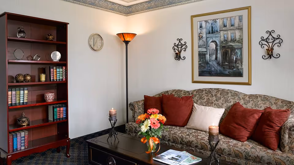 A cozy living room area with a floral patterned sofa adorned with red and beige cushions. In front of the sofa is a dark wooden coffee table with a vase of colorful flowers and two lit candles. To the left, there is a tall wooden bookshelf filled with books and decorative items. A floor lamp with an amber shade stands in the corner, and a framed painting of a street scene hangs on the wall above the sofa, flanked by two decorative wall sconces. A round wall clock is mounted on the adjacent wall.