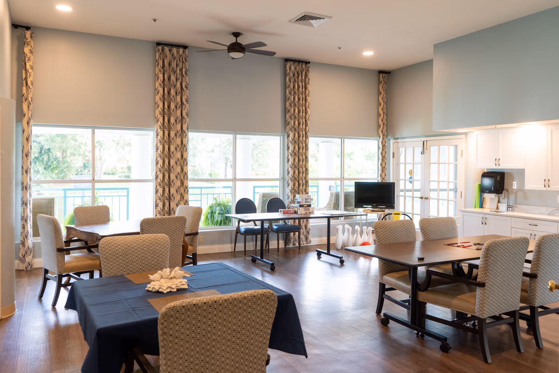 A bright and spacious common room with several tables and cushioned chairs arranged for group activities. Large windows with patterned curtains let in natural light. A ceiling fan is mounted above, and a small TV is placed near a set of bowling pins and balls. The room also features a kitchenette area with white cabinets and a sink.
