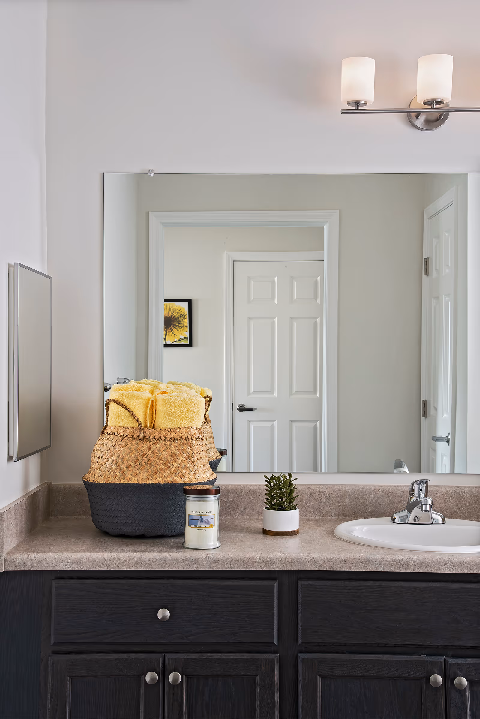 A bathroom countertop with a sink, a small potted plant, a candle, and a woven basket filled with yellow towels. Above the countertop is a large mirror reflecting a white door and a framed picture on the opposite wall. A two-light wall fixture is mounted above the mirror.