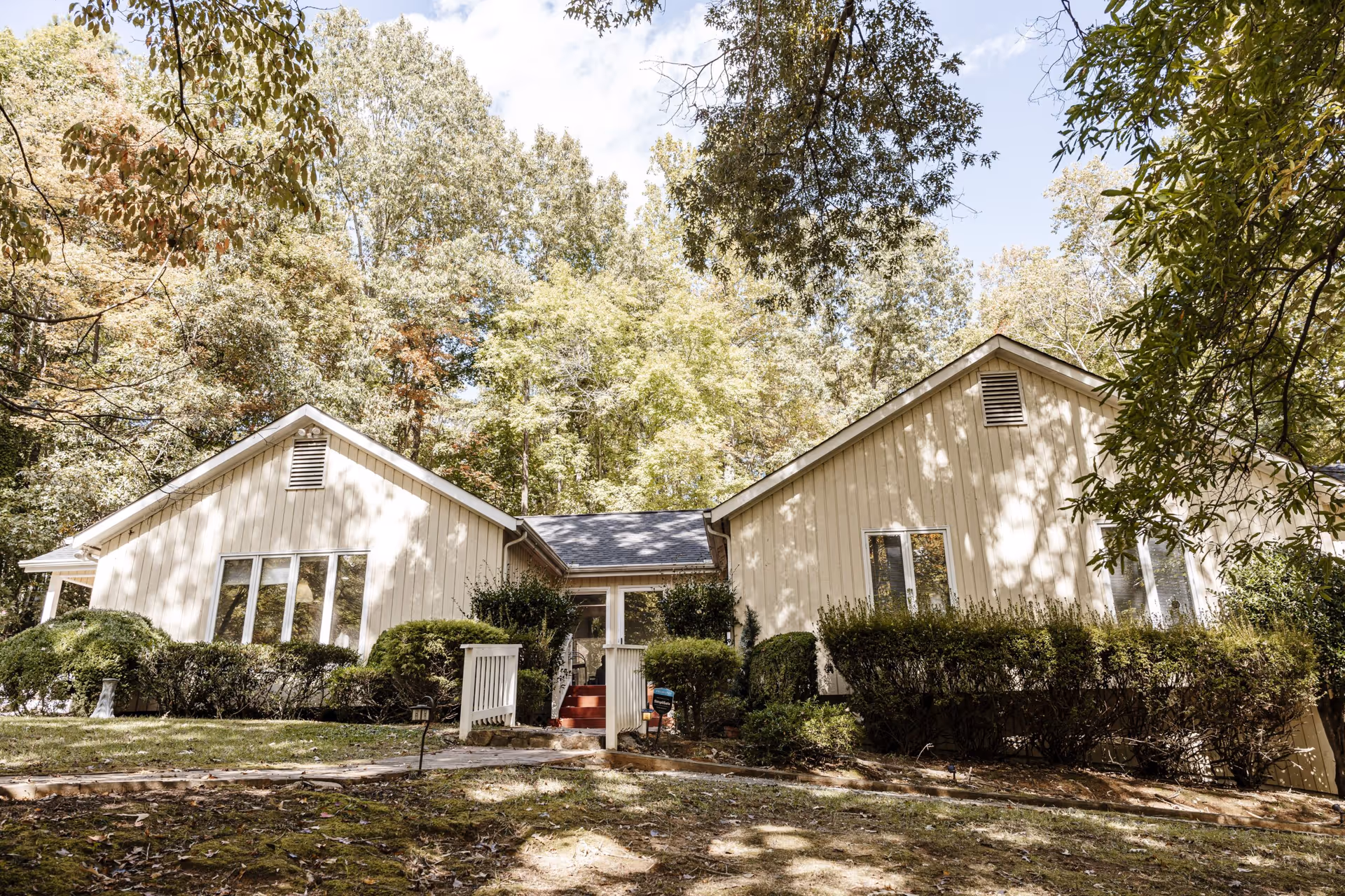 Exterior view of a beige single-story building with large windows surrounded by bushes and trees with sunlight filtering through the leaves.