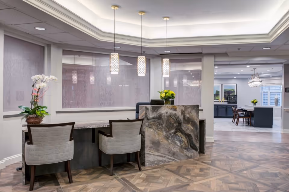 Reception area with a marble front desk and two upholstered chairs. Three pendant lights hang from the ceiling above the desk. A potted orchid and a vase with yellow flowers decorate the desk. In the background, there is a dining area with tables, chairs, and framed artwork on the wall.