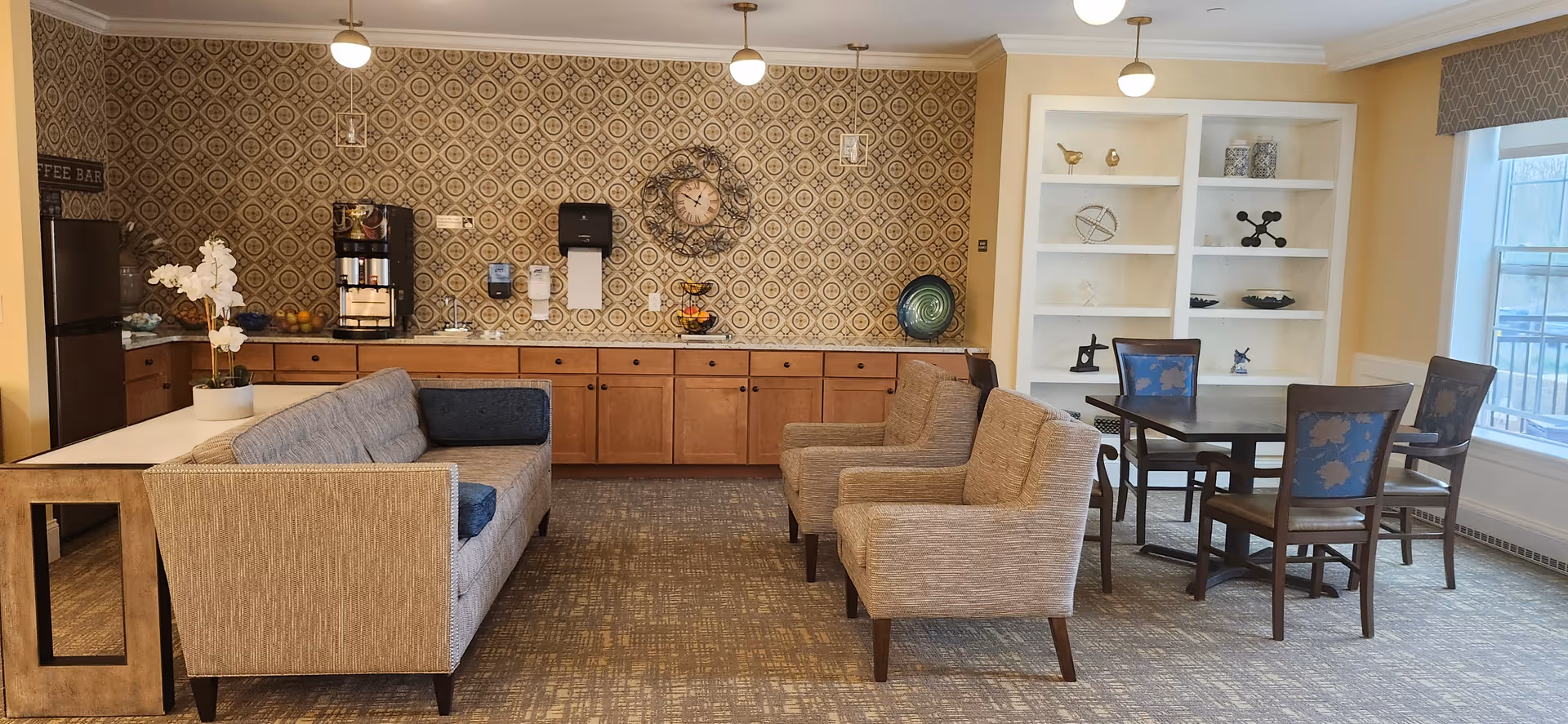 A cozy common area with a patterned wallpapered wall featuring a clock and a coffee station. The room includes a beige sofa, two beige armchairs, a wooden table with four chairs near a window, and built-in white shelves with decorative items.