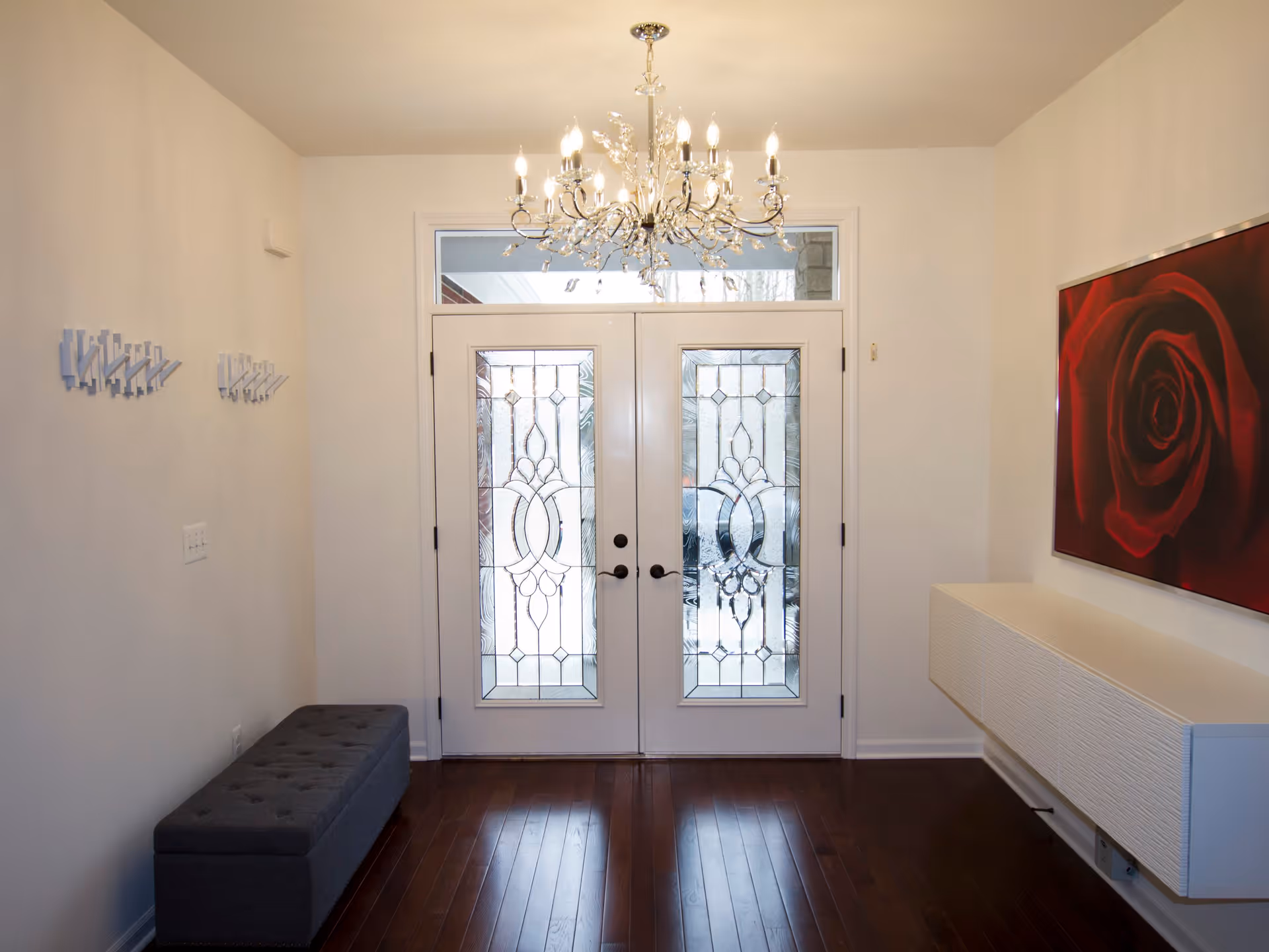 Entryway with double glass doors featuring decorative frosted glass panels, a crystal chandelier hanging from the ceiling, a gray cushioned bench on the left, white wall-mounted coat hooks, a white floating cabinet on the right, and a large red rose painting above the cabinet.