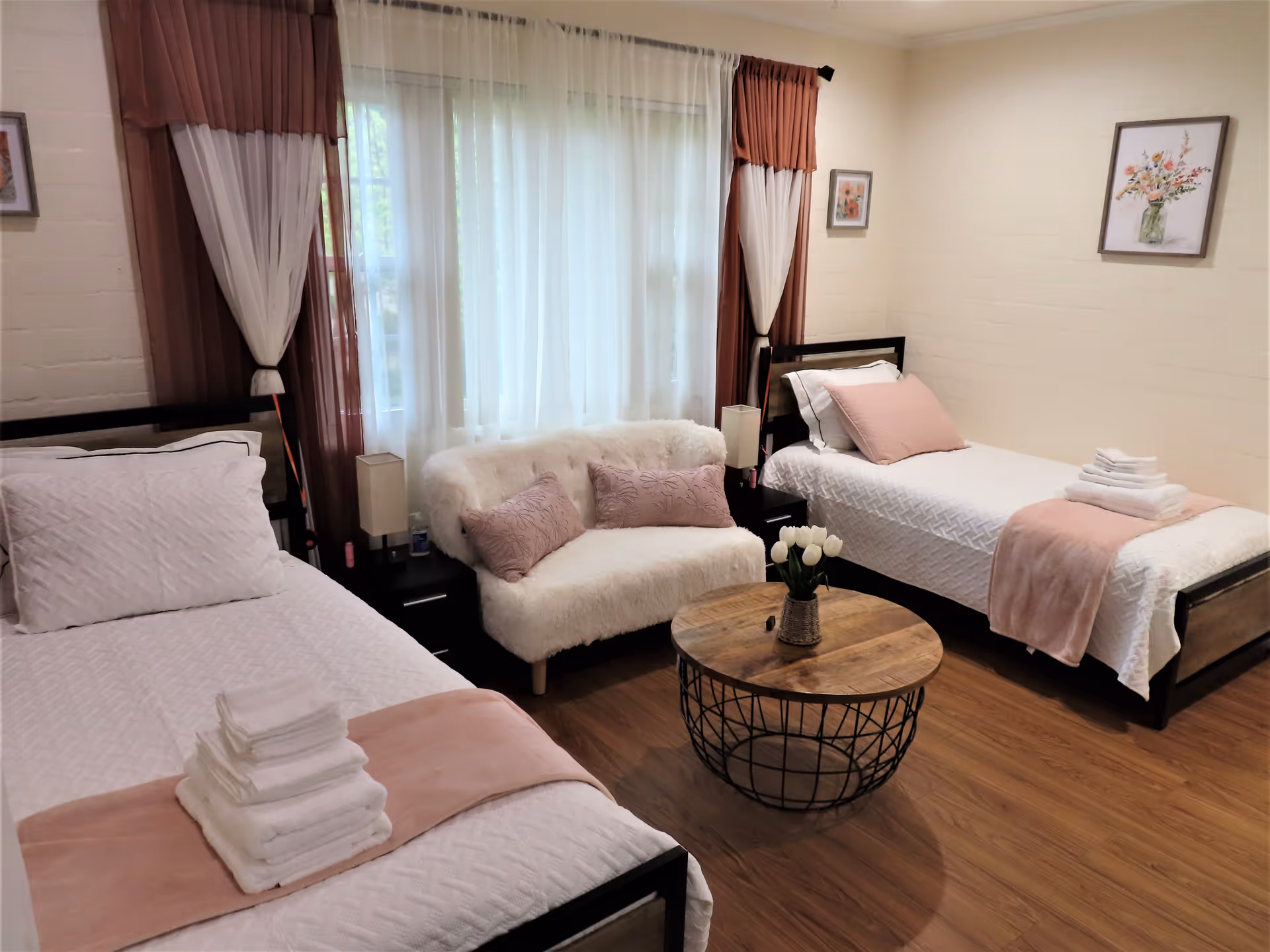 Bright shared bedroom with two twin beds, a small white loveseat, round wooden coffee table, and soft pink accents.
