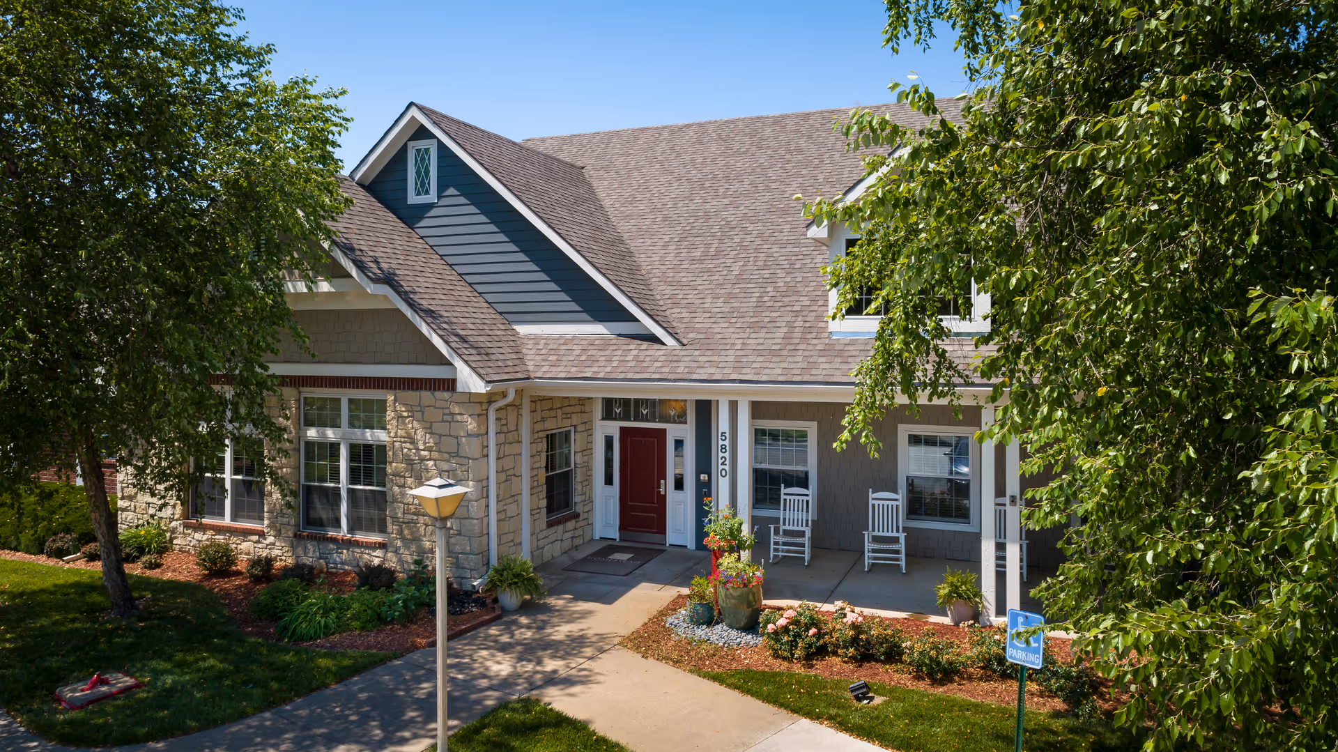 Front exterior view of a single-story assisted living facility with a stone and siding facade, a covered porch with two white rocking chairs, potted plants, and a walkway leading to a red front door. The building is surrounded by green trees and landscaping under a clear blue sky.
