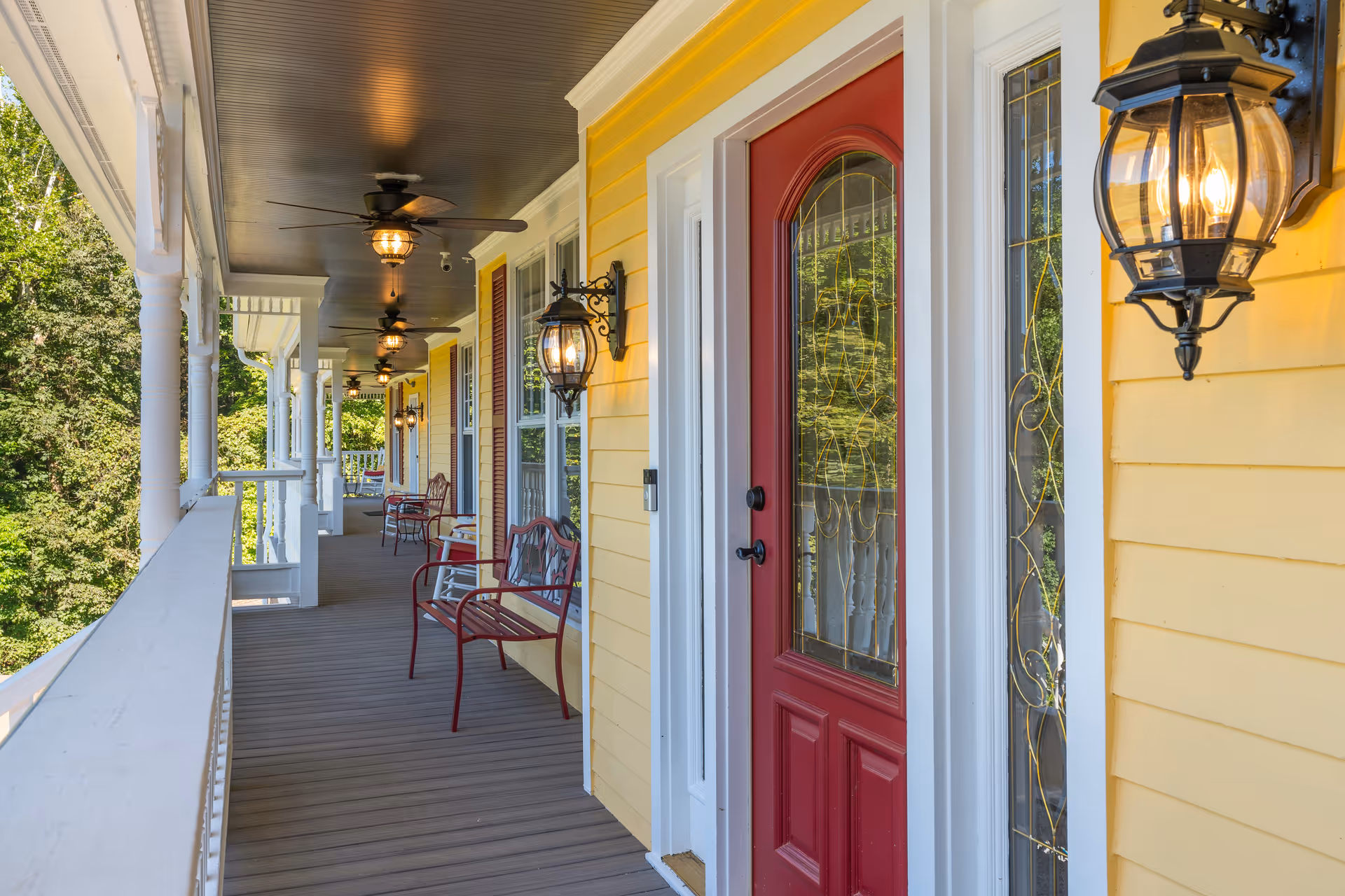 Covered yellow-painted porch with red entry doors, hanging lanterns, ceiling fans, and benches along a railing overlooking trees.