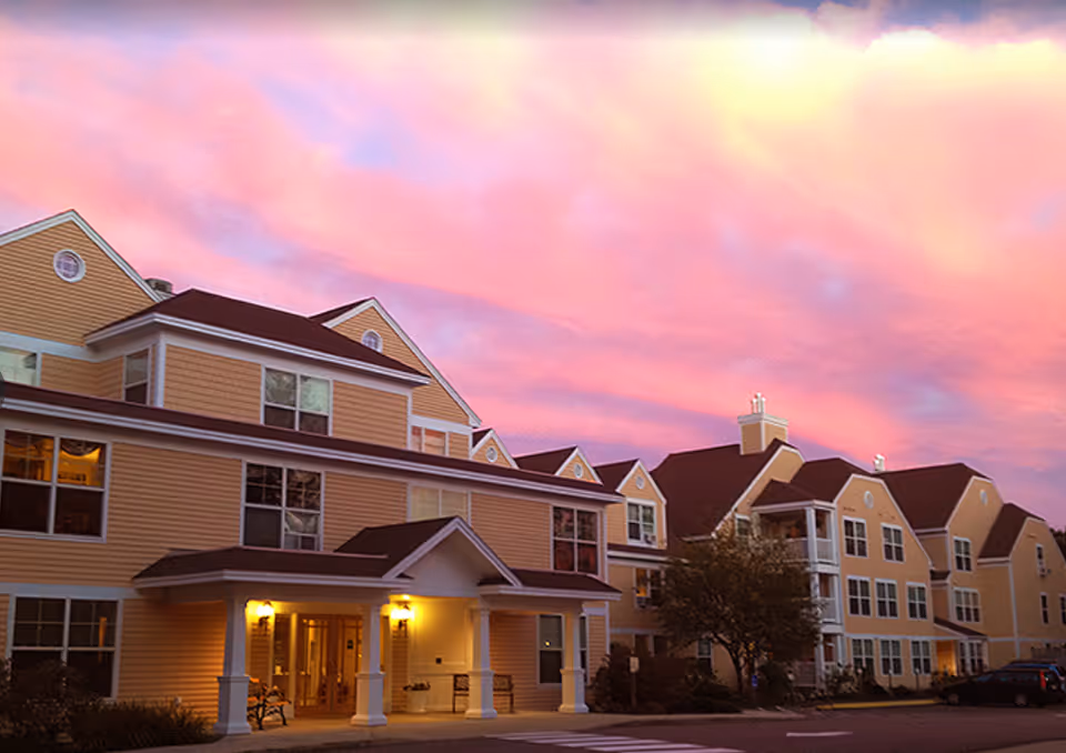 Front exterior of a multi-story yellow senior living building under a pink sunset sky.