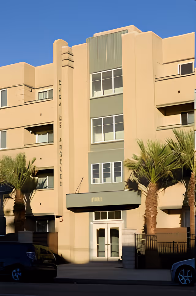 Exterior view of a multi-story beige building with several windows and palm trees in front, under a clear blue sky.