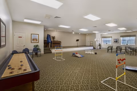 A spacious activity room with a patterned carpet, featuring a shuffleboard table on the left, a small stage with a piano and microphone stand at the back, and various game setups including bowling pins, cornhole boards, and ladder toss. Several chairs are arranged along the walls, and framed pictures hang on the walls under bright ceiling lights.