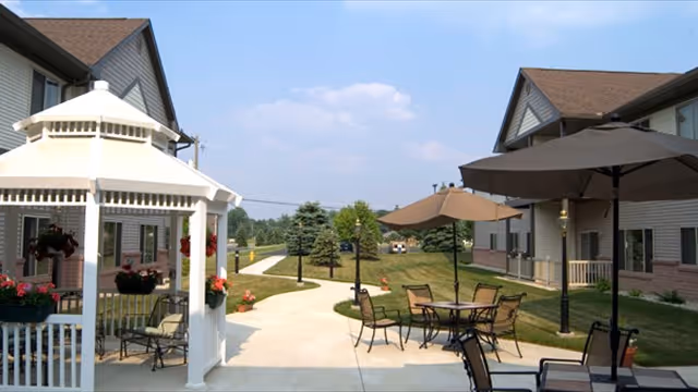 Outdoor courtyard with a white gazebo, patio tables and umbrellas, paved walkways, lawn and surrounding two-story residential buildings.