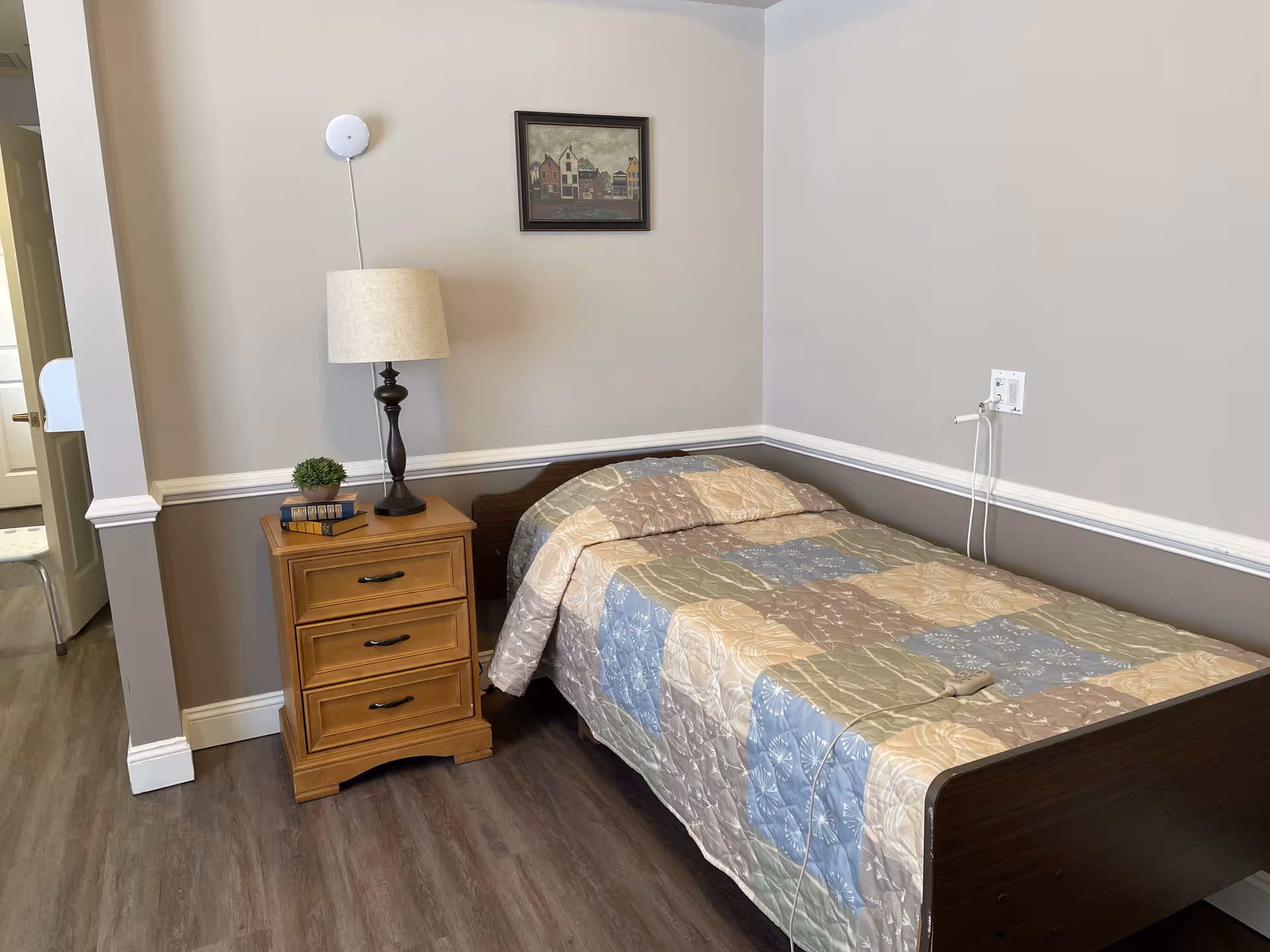 A small bedroom with a single bed covered with a patchwork quilt in shades of blue, beige, and green. Next to the bed is a wooden nightstand with three drawers, a table lamp, two books, and a small potted plant. A framed picture of houses hangs on the wall above the nightstand. The room has light gray walls with a white chair rail and dark wood flooring.