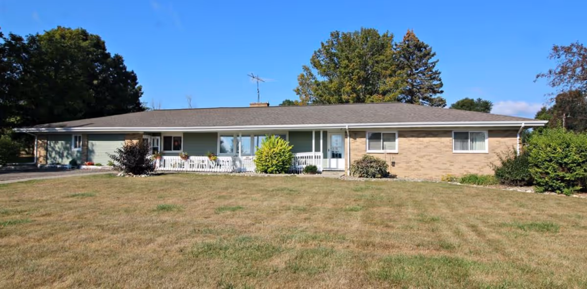 Single-story building with a combination of green siding and brick exterior, a white porch railing, and several windows. The building is surrounded by a large grassy lawn with some bushes and trees in the background under a clear blue sky.