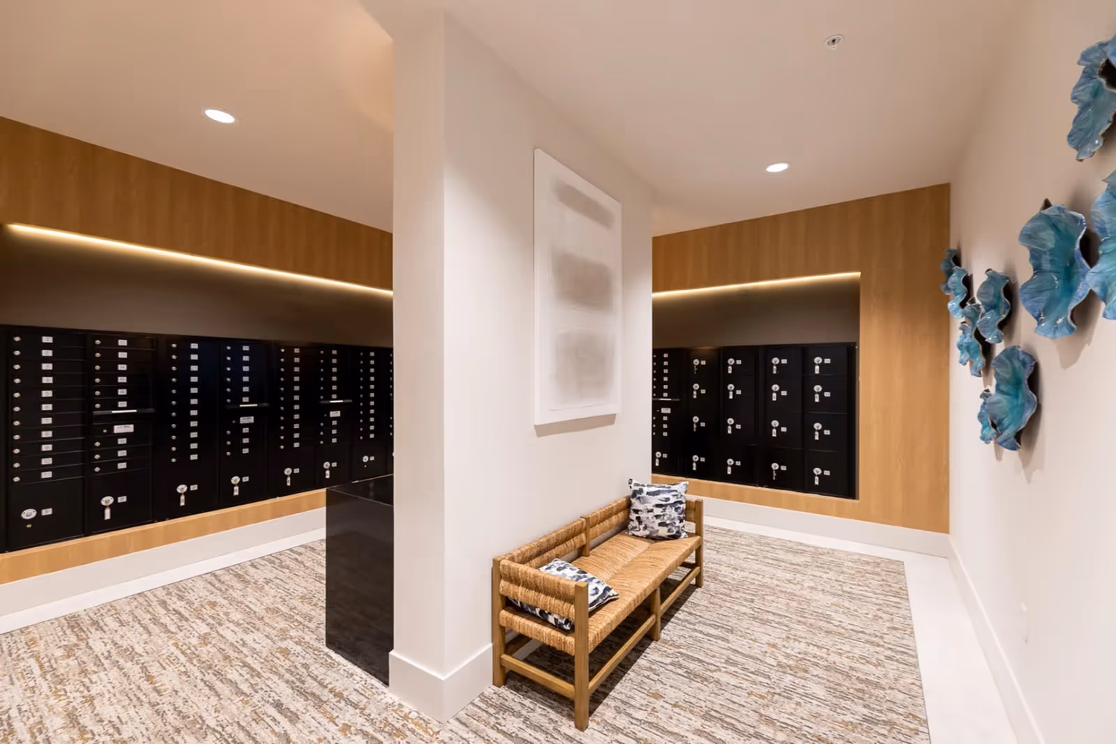 Interior view of a mailroom area with black mailboxes mounted on wooden panels along two walls. A wooden bench with woven seating and two patterned cushions is placed against a white wall in the center. The floor is covered with a textured carpet, and blue decorative wall art is visible on the right wall.