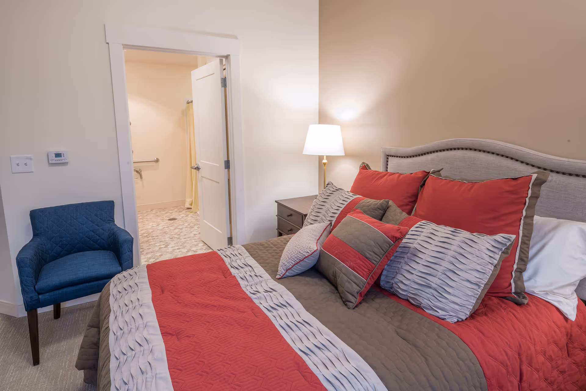 Well-lit bedroom with a neatly made bed with red and gray pillows, a blue chair, and an open door leading to a bathroom.