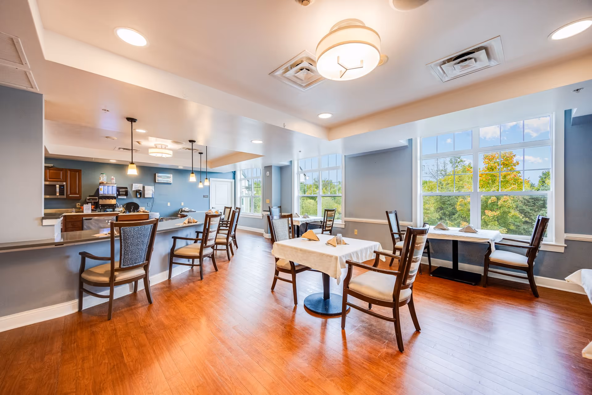 A bright dining area with wooden floors, several tables covered with white tablecloths and napkins, and wooden chairs. Large windows let in natural light and offer a view of green trees outside. The room also features a counter with bar stools and pendant lighting above it.