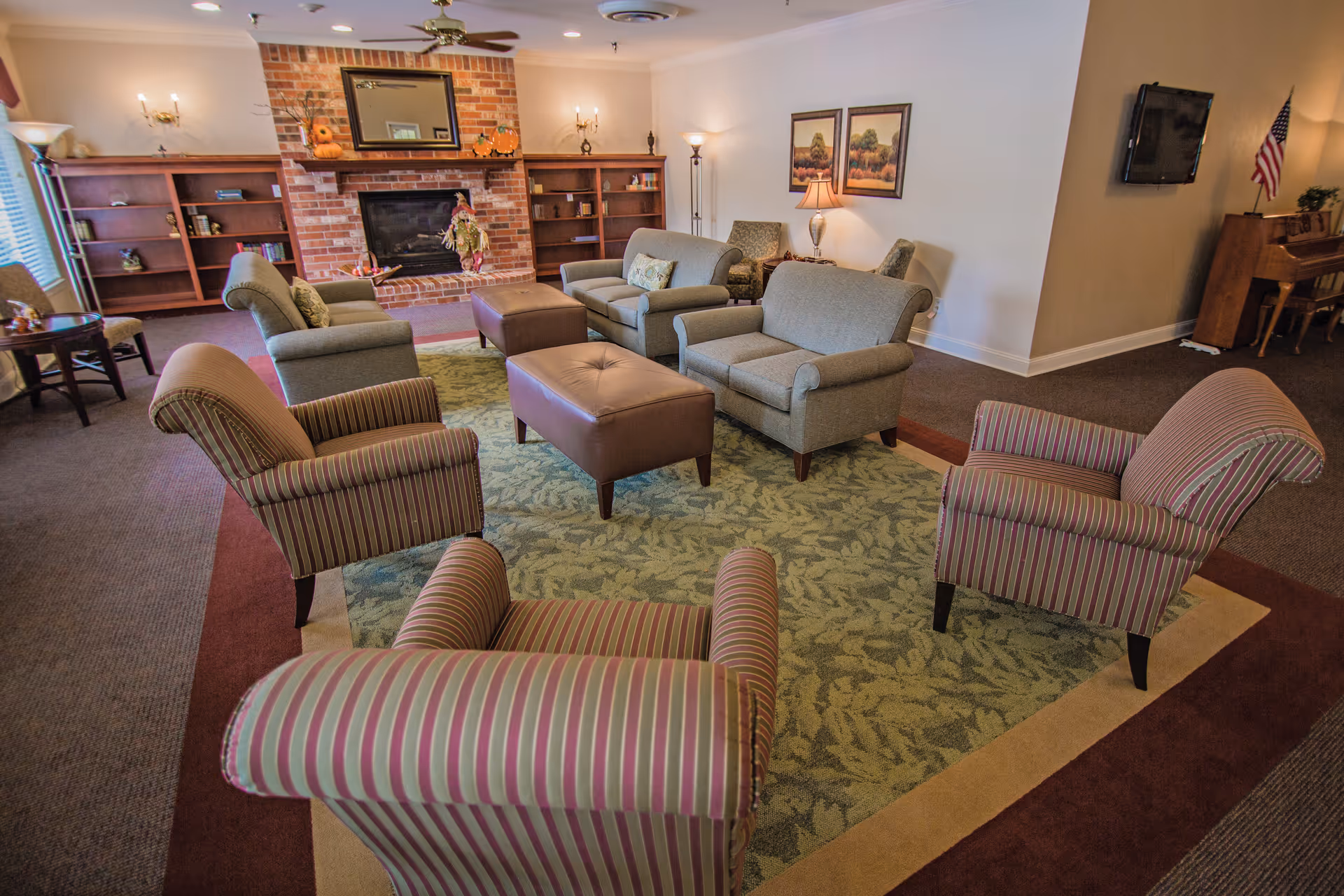 A cozy living room area with several upholstered armchairs and sofas arranged around two brown ottomans on a patterned area rug. The room features a brick fireplace with a mirror above it, built-in wooden shelves on either side, and various lamps providing warm lighting. A piano and an American flag are visible in the background.