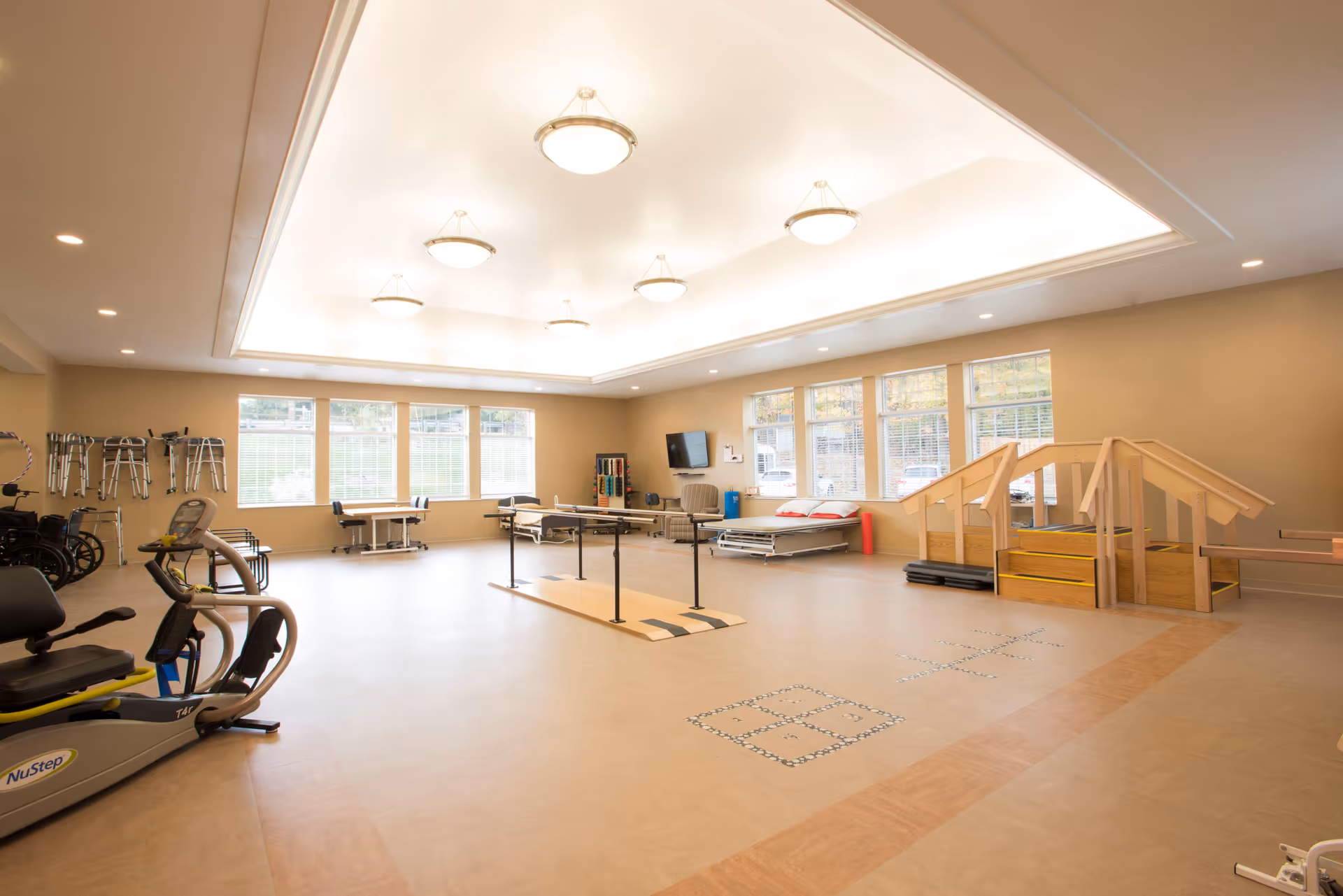 A spacious rehabilitation room with exercise equipment, parallel bars for walking practice, a set of wooden stairs with handrails for physical therapy, wheelchairs and walkers lined up against the wall, and large windows letting in natural light.