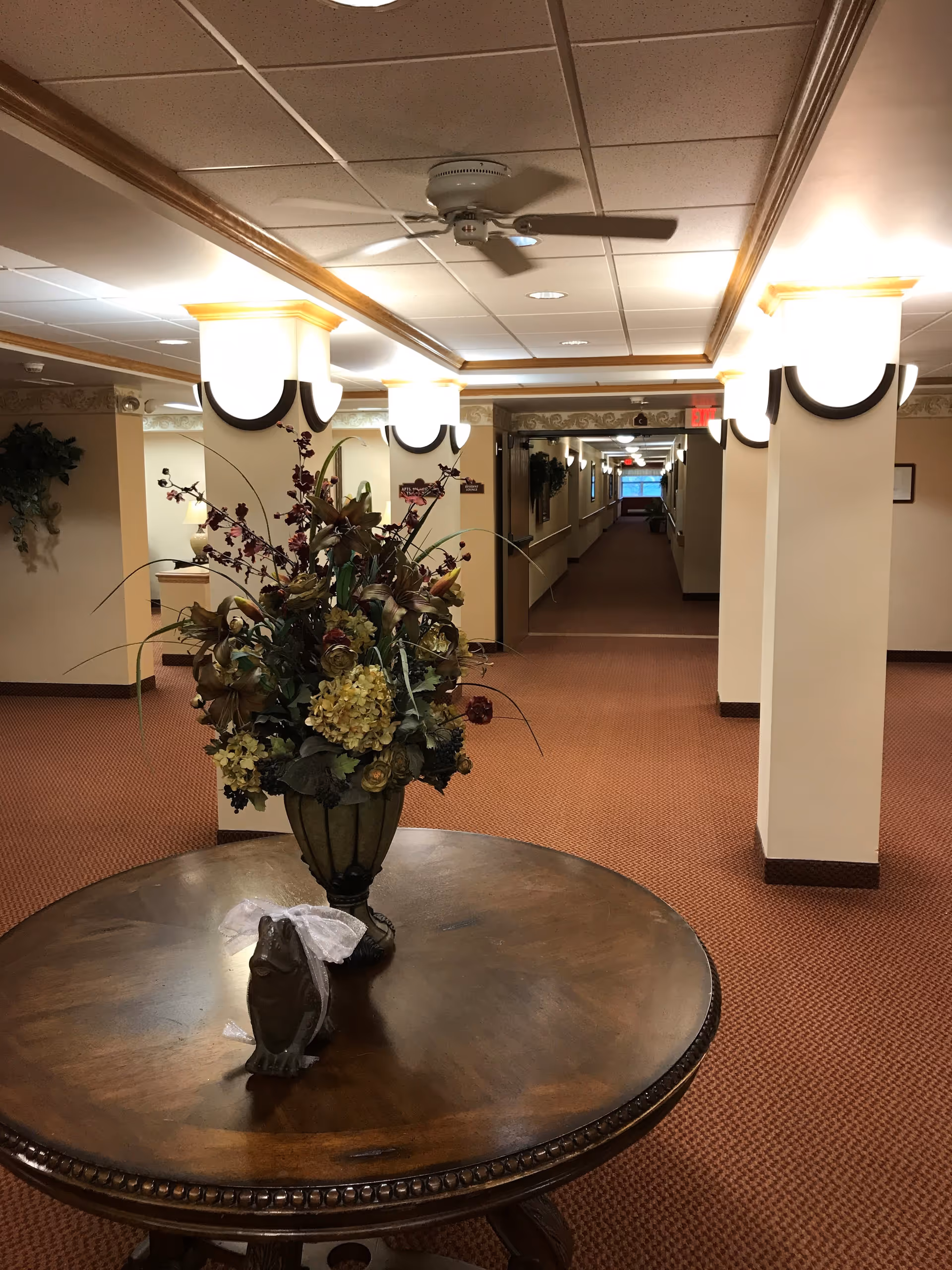 Interior hallway of a senior living facility with beige walls, carpeted floor, and ceiling lights. A round wooden table with a decorative flower arrangement and a small frog figurine with a white bow is in the foreground. The hallway extends into the distance with handrails along the walls and an exit sign visible at the far end.