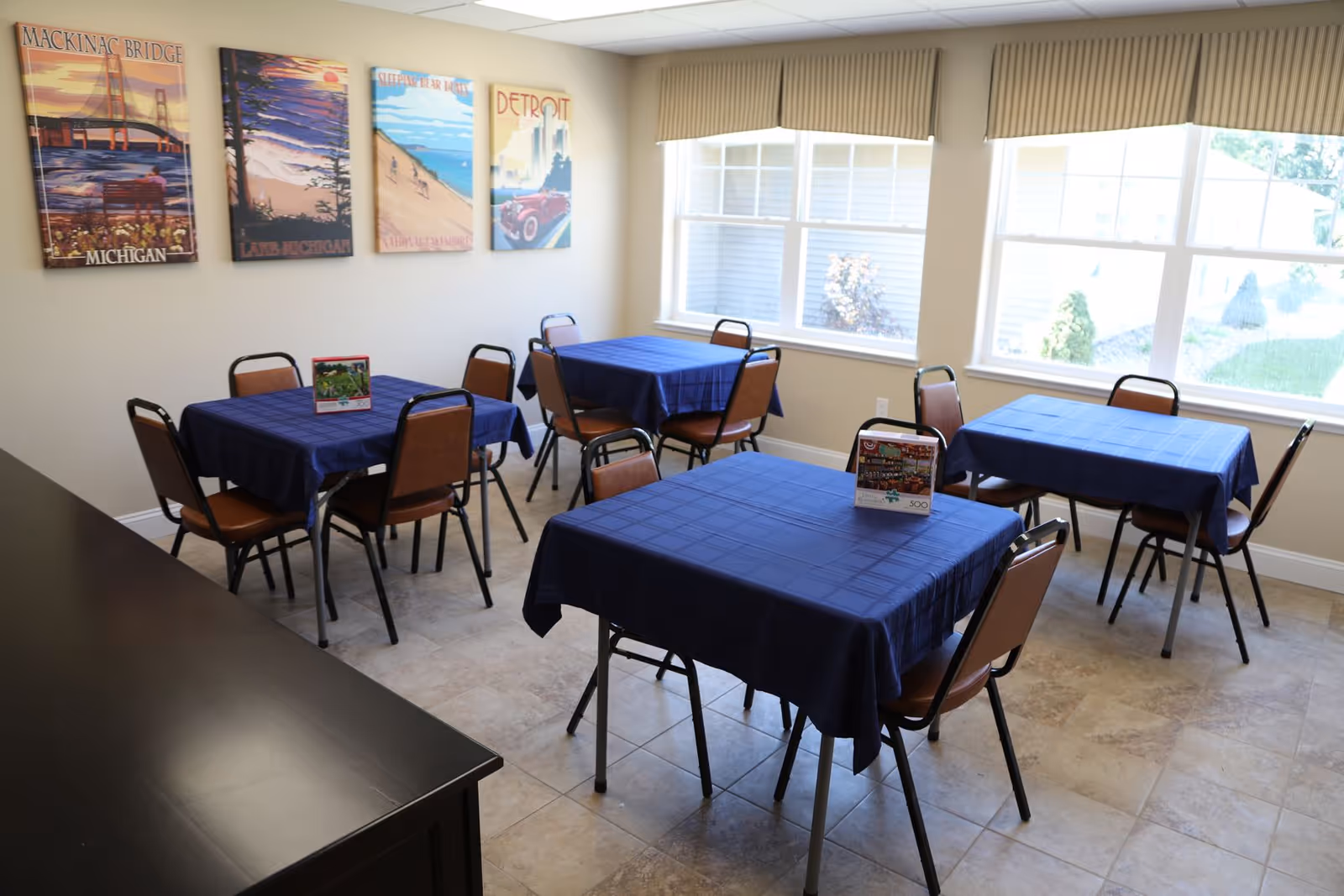A small dining room with four square tables covered with blue tablecloths, each surrounded by four brown chairs. The room has large windows with beige valances letting in natural light. On the wall are four colorful posters depicting Michigan landmarks and scenes, including Mackinac Bridge, Lake Michigan, Sleeping Bear Dunes, and Detroit. The floor is tiled, and there is a dark wooden cabinet or counter visible in the foreground.